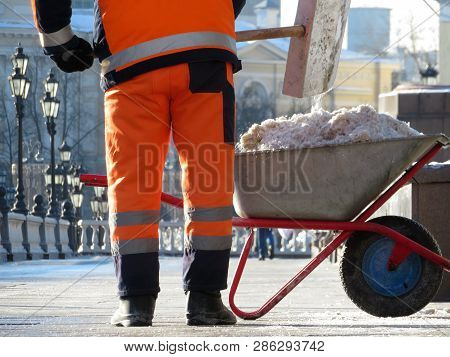 Snow Removal In City, Municipal Worker With Shovel Loading Snow In The Wheelbarrow. Clearing Snow In