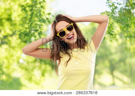 summer, accessory and people concept - smiling young woman or teenage girl in yellow t-shirt and sunglasses holding to her head over green natural background