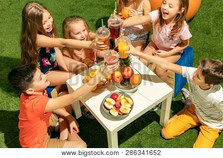 Kids Fashion Concept. The Group Of Teen Boys And Girls Sitting On Green Grass At Park. Children Colo