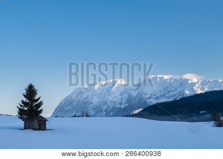 Snow Covered Small Hut With A Tree On A Field With Mountains Grimming, Schartenspitze And Steinfelds