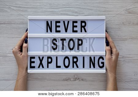 Female Hands Hold Modern Board With 'never Stop Exploring' Word Over White Wooden Surface, Top View.