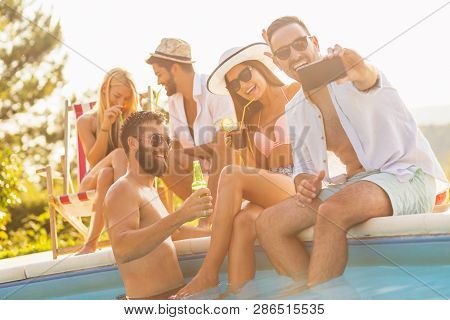 Group Of Friends Having Fun At A Poolside Summer Party, Sitting At The Edge Of A Swimming Pool, Drin
