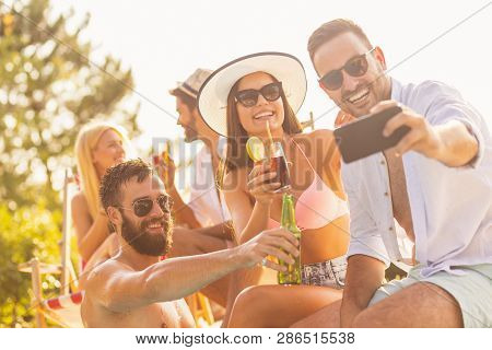 Group Of Friends Having Fun At A Poolside Summer Party, Sitting At The Edge Of A Swimming Pool, Drin