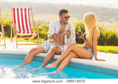 Couple In Love At A Poolside Summer Party, Sitting At The Edge Of A Swimming Pool, Drinking Beer And