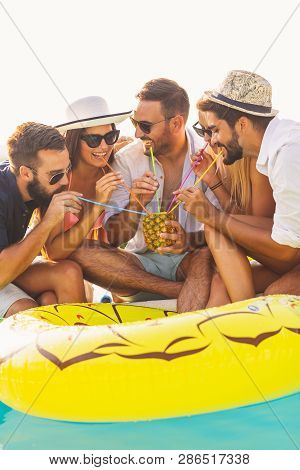 Group Of Friends At A Poolside Summer Party, Sitting At The Edge Of A Swimming Pool, Drinking A Pine