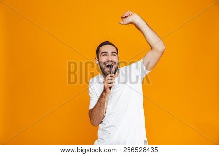 Photo of optimistic man 30s listening to music using earphones and mobile phone isolated over yellow background