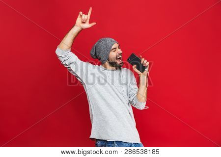 Image of optimistic man 30s singing while listening to music with earphones and mobile phone isolated over red background
