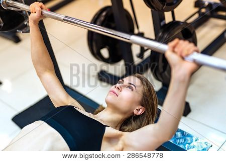 Athletic Girl Raises Barbell Lying On The Bench On The Simulator In The Gym. Young Woman Doing Inten
