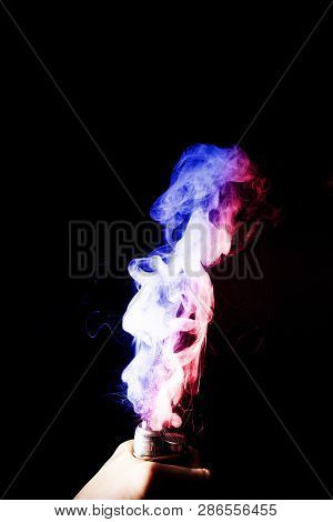 Male Hand Holds Vape. Vape Clouds Splash From Vape Coil On Black Background. Fog Is Dual Color -- Bl