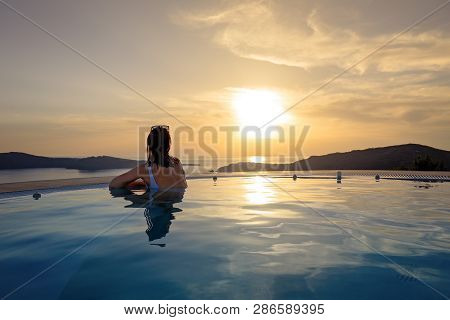 Woman In Infinity Swimming Pool At Golden Sunset. Girl Admiring Sunset From The Pool. Summer Vacatio