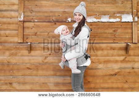 Little Baby Girl And Her Mother Babywearing The Ergo Carrier In Winter Wooden Background