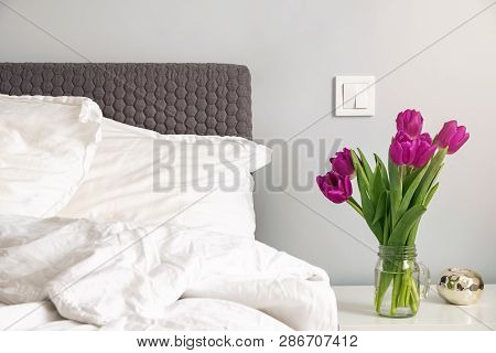 Purple Tulips Standing On A Nightstand In The Modern White Bedroom Near The Bed.