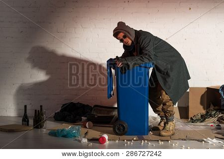 Homeless Man In Dark Clothes Rummaging In Trash Container