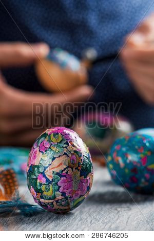 closeup of a young cacausian man decorating some homemade easter eggs by gluing some pieces of different papers, on a gray rustic wooden table