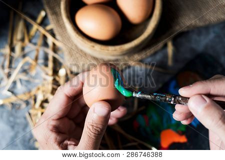 closeup of a young cacausian man painting an easter egg, on a gray rustic wooden table, next to some brown chicken eggs in a boxwood bowl and some straw stalks