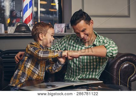 Young Father And His Stylish Little Son In The Barbershop In The Waiting Room.
