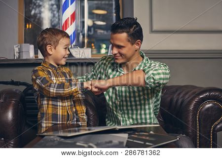 Young Father And His Stylish Little Son In The Barbershop In The Waiting Room.