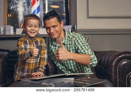 Young Father And His Stylish Little Son In The Barbershop In The Waiting Room.