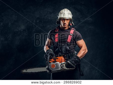 Young Logger Wearing Protective Clothing And Helmet Posing With A Chainsaw In A Dark Studio And Look