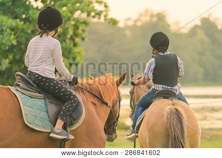 Kids Learn To Ride A Horse Near The River Before Sunset.