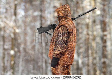 Hunter In Autumn Camouflage With A Gun On His Shoulder In The Winter Forest