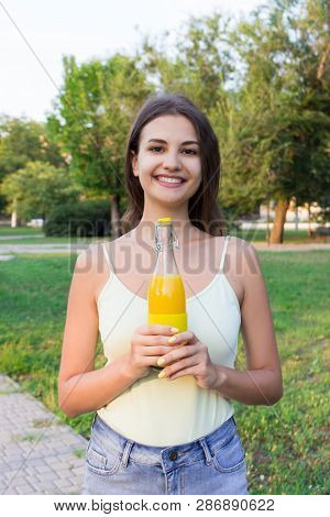 Cute Girl Is Having A Walk In The Park Holding A Bottle Of Orange Juice In A Sunny Wam Day