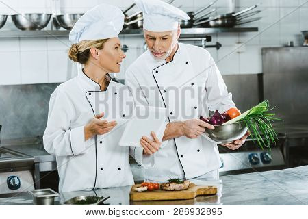 Female And Male Chefs In Iniforms Using Digital Tablet During Cooking In Restaurant Kitchen