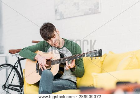 Handsome Young Man Playing Acoustic Guitar While Sitting On Yellow Sofa