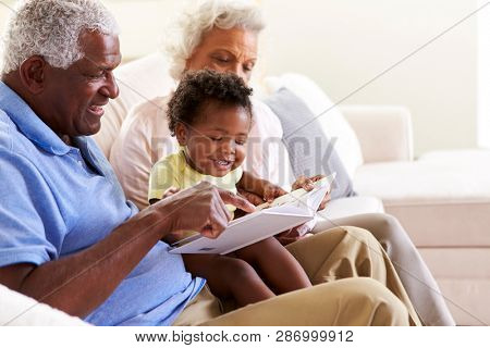 Grandparents Sitting On Sofa At Home With Baby Granddaughter Reading Book Together