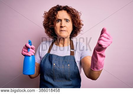 Middle age curly hair woman cleaning doing housework wearing apron and gloves using spayer annoyed and frustrated shouting with anger, crazy and yelling with raised hand, anger concept