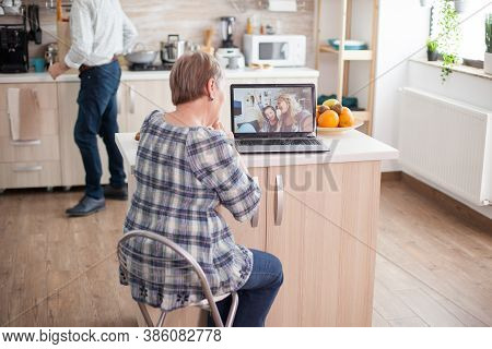 Happy Senior Woman During A Video Conference With Family Using Laptop In Kitchen. Online Call With D