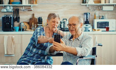 Smiling Senior Woman And Her Disabled Husband In Wheelchair Using Smartphone In Kitchen. Paralysied 