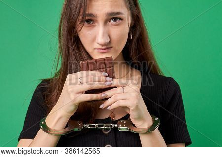 Girl In Handcuffs Is Holding Chocolate. Breaking Diet Concept. Close-up Portrait Of Young Woman With