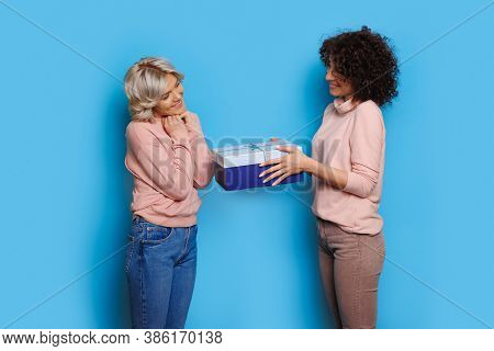 Brunette Woman With Curly Hair Giving A Gift To Her Blonde Sister Who Is Gesturing Happiness On A Bl