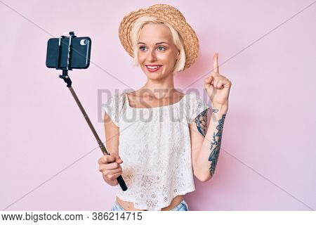 Young blonde woman with tattoo taking a selfie photo with smartphone smiling with an idea or question pointing finger with happy face, number one 