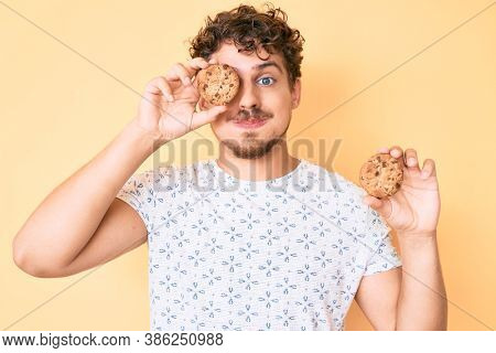 Young caucasian man with curly hair holding cookie puffing cheeks with funny face. mouth inflated with air, catching air. 