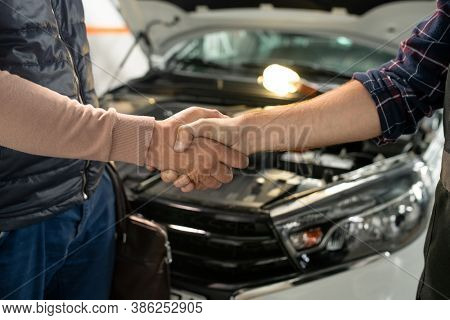 Young contemporary technician of car service shaking hand of male client against front part of broken car with open engine compartment
