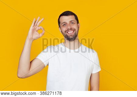 Young Man With A Beard In A White T-shirt Happy Face, Smiling Positively, Makes Okay Sign With Hand 