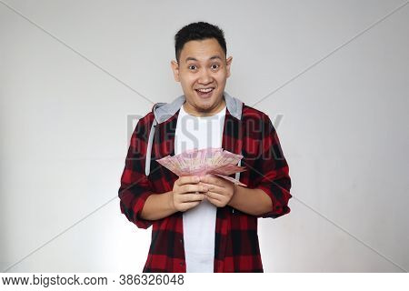 Portrait Of Happy Young Indonesian Man Holding Rupiah Money, Smiling Laughing Winning Gesture, Over 