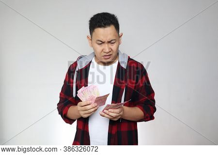 Confused Stressed Asian Man Holding Indonesian Rupiah Cash In Hand, Thinking Wondering How To Spend.