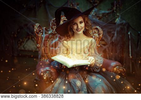 Portrait of a charming woman witch in a hat and beautiful dress sitting in an armchair with a book of spell in the old castle. Halloween. Celebration.