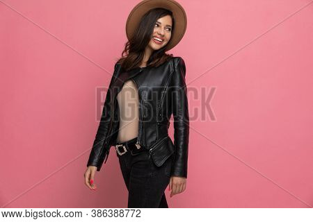 Cheerful fashion model laughing and looking back over shoulder and wearing hat while standing on pink studio background