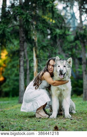 Young Woman With Innate Disorder Dwarfism Embraces Malamute Dog While Walking In Park.