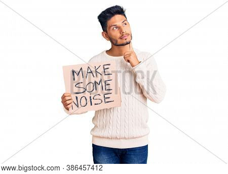 Handsome latin american young man holding make some noise banner serious face thinking about question with hand on chin, thoughtful about confusing idea 