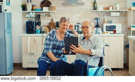 Senior Couple Laughing And Waving During A Video Call With Grandchildrens Using Tablet Computer In K