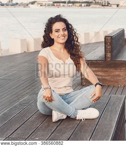 Young Women Wearing T-shirt And Jeans Sits On The Wooden  Bench And Smile
