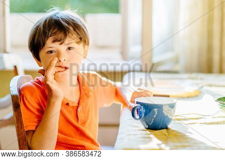 Thoughtful Child Boy Wearing Orange T-shirt With Down Syndrome Sitting At Table At Home And Bite Nai