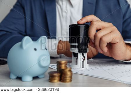 Businessman Is Stamping Important Contract In Office. Blue Piggy Bank With Stack Of Coins On Desk. E