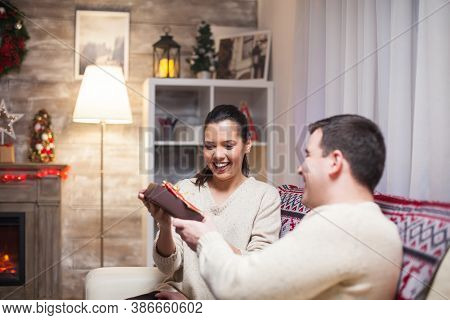 Excited Young Woman While Her Husband Is Giving Her A Gift While Celebrating Christmas.