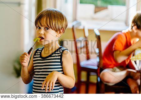 Cute Little Preschooler Boy In Striped T-shirt Help His Mother At Kitchen In Home, Child Eat Slice O